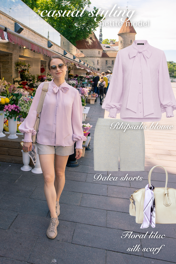 Woman wearing a premium lavender silk neck tie blouse and gray tweed shorts with a floral lilac silk scarf, standing on a street with flower market stalls.