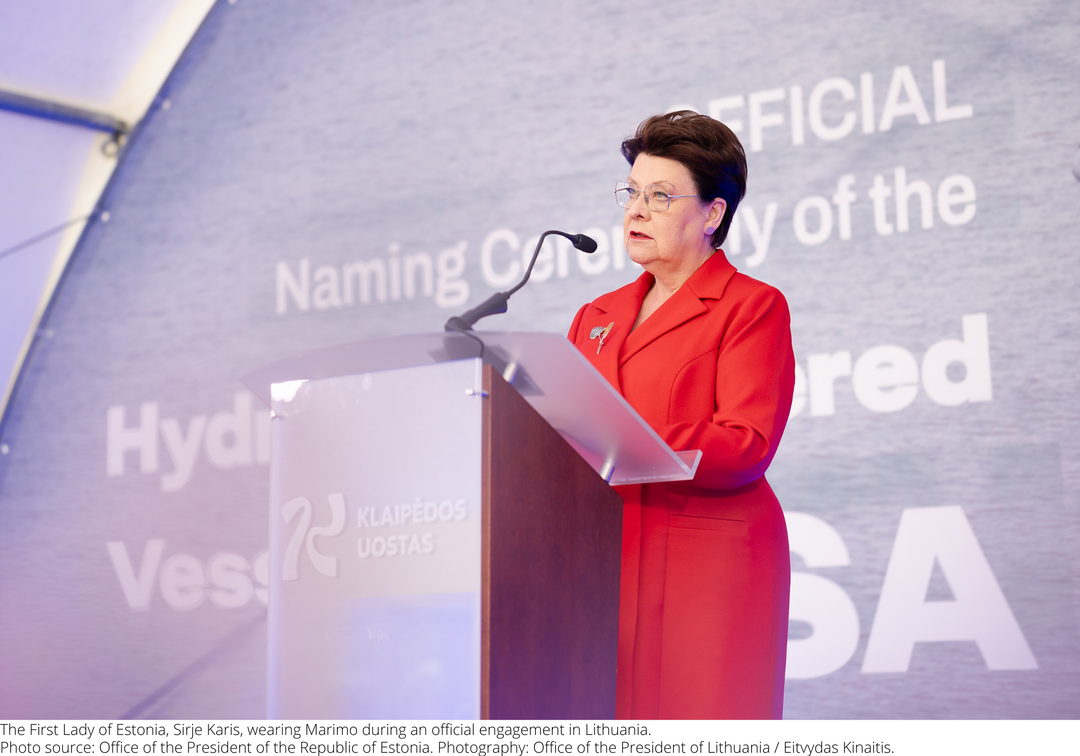 The first lady of Estonia in a bright red suit stands behind a clear podium with a microphone, speaking at an event. 