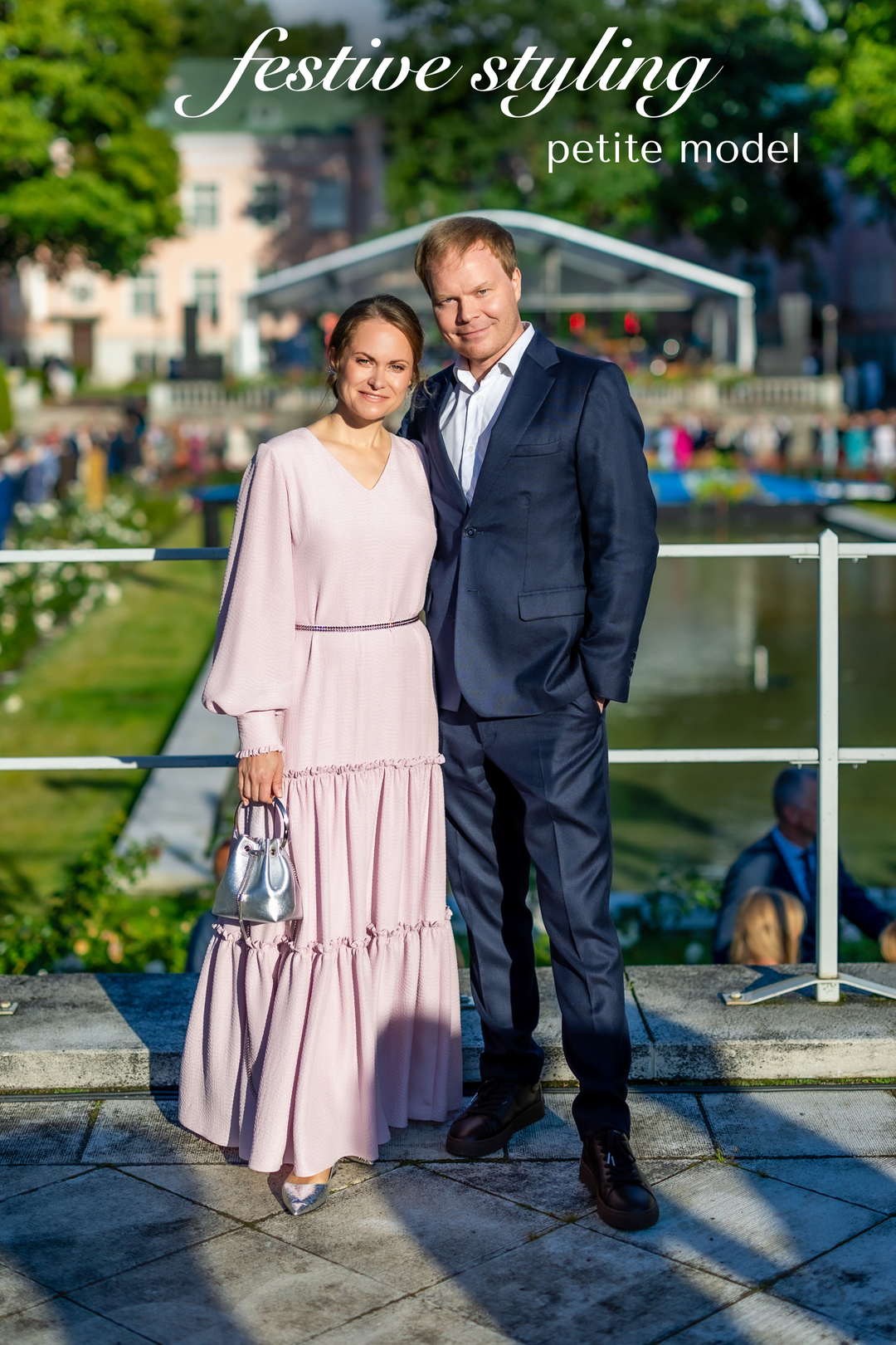 A couple posing outdoors by a canal, with the woman wearing a light pink tiered maxi dress and the man wearing a navy blue suit. The text 'festive styling petite model' is written at the top.