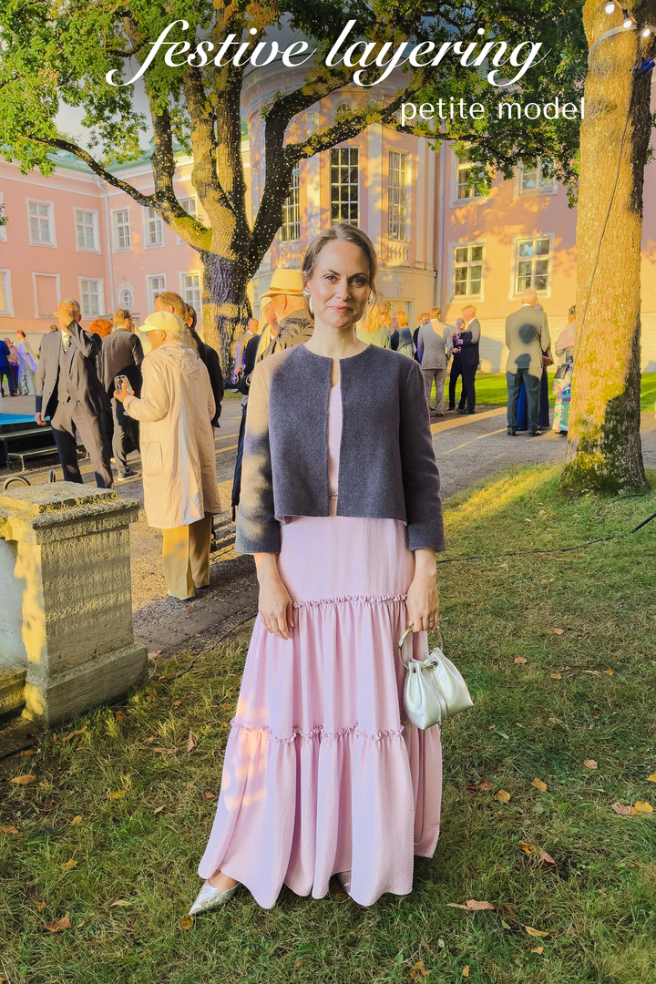 A woman stands outdoors on grass in front of a building and trees, wearing a gray cropped wool coat over a light pink tiered maxi skirt. She is holding a small white handbag.