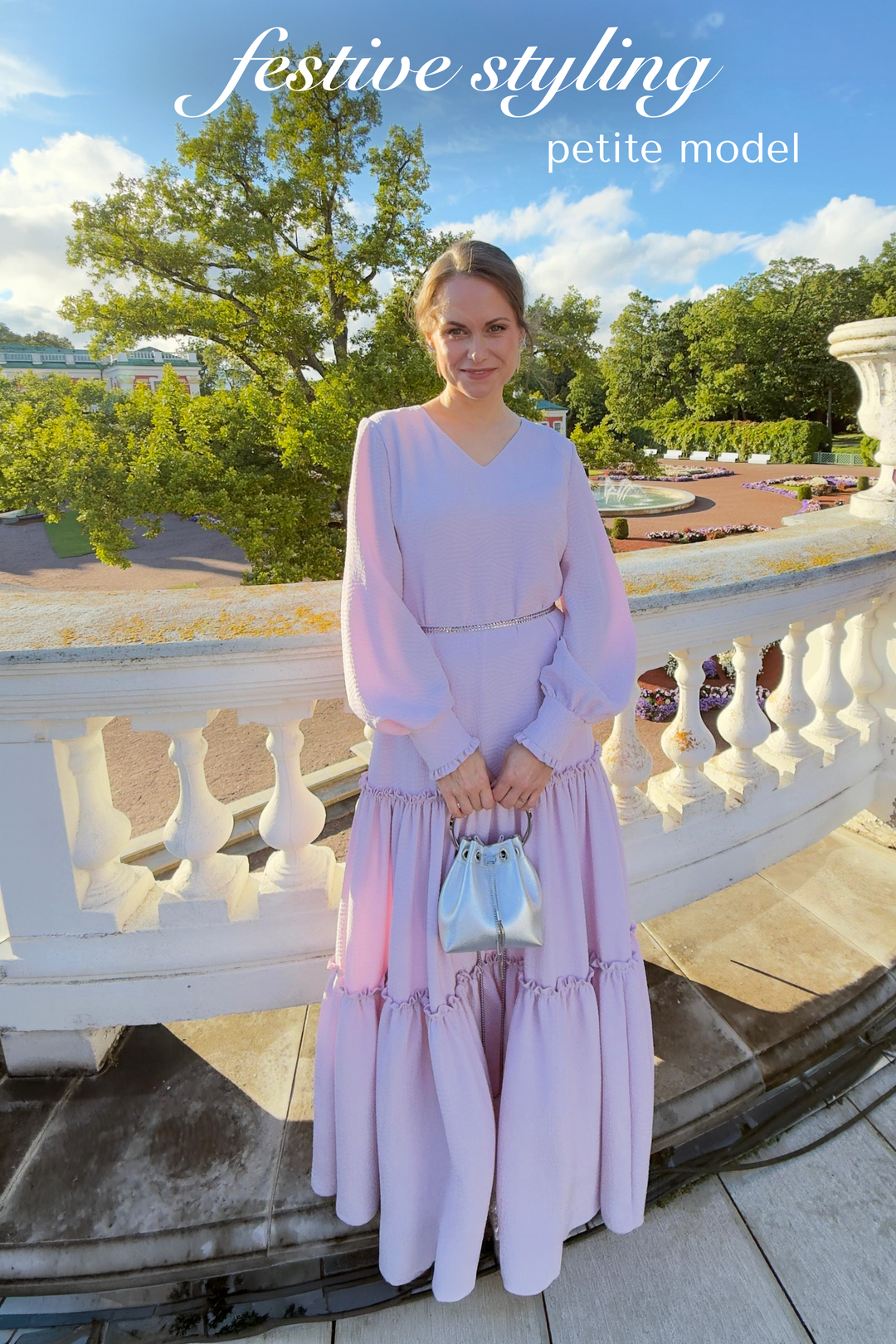 A woman stands outdoors on a stone terrace wearing a long lavender dress with long sleeves and a tiered skirt, holding a small light blue handbag. Trees, a white balustrade, and blue sky are visible in the background.