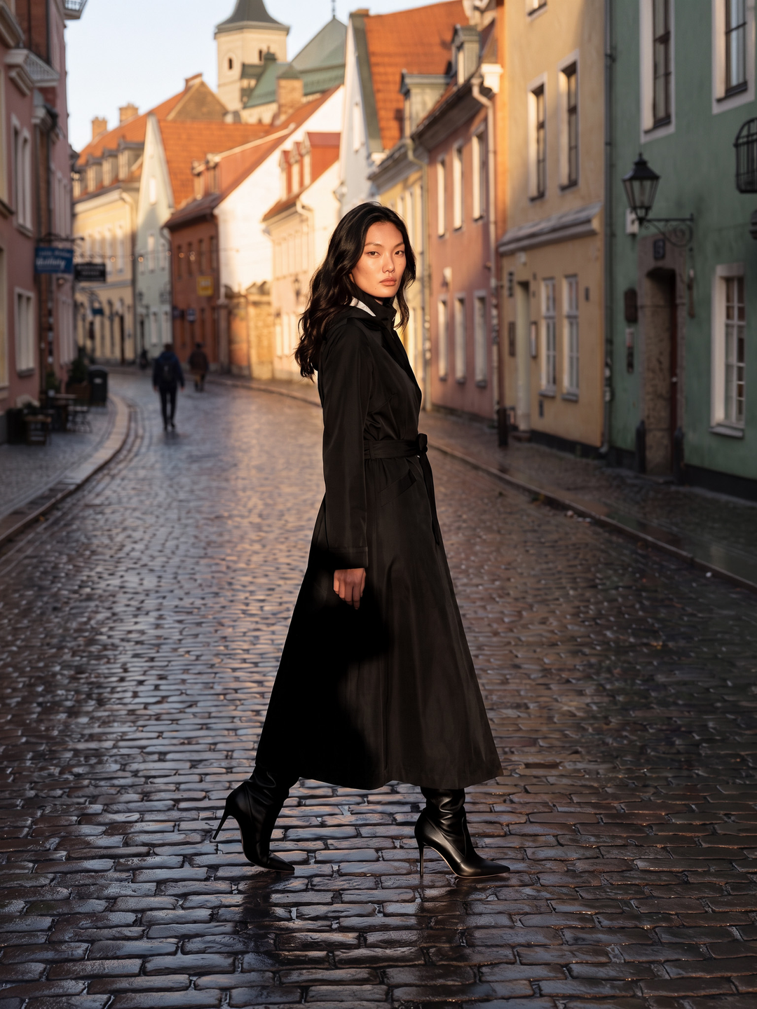 A woman wearing a long black coat and black high-heeled boots walks along a cobblestone street lined with colorful historic buildings.