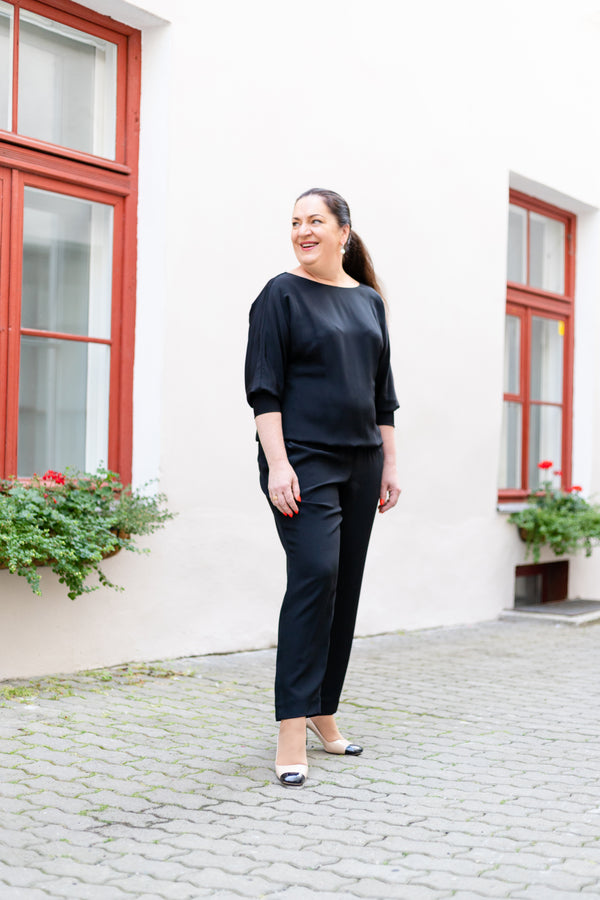 Woman in a black jumpsuit standing in front of a white building with red window frames.
