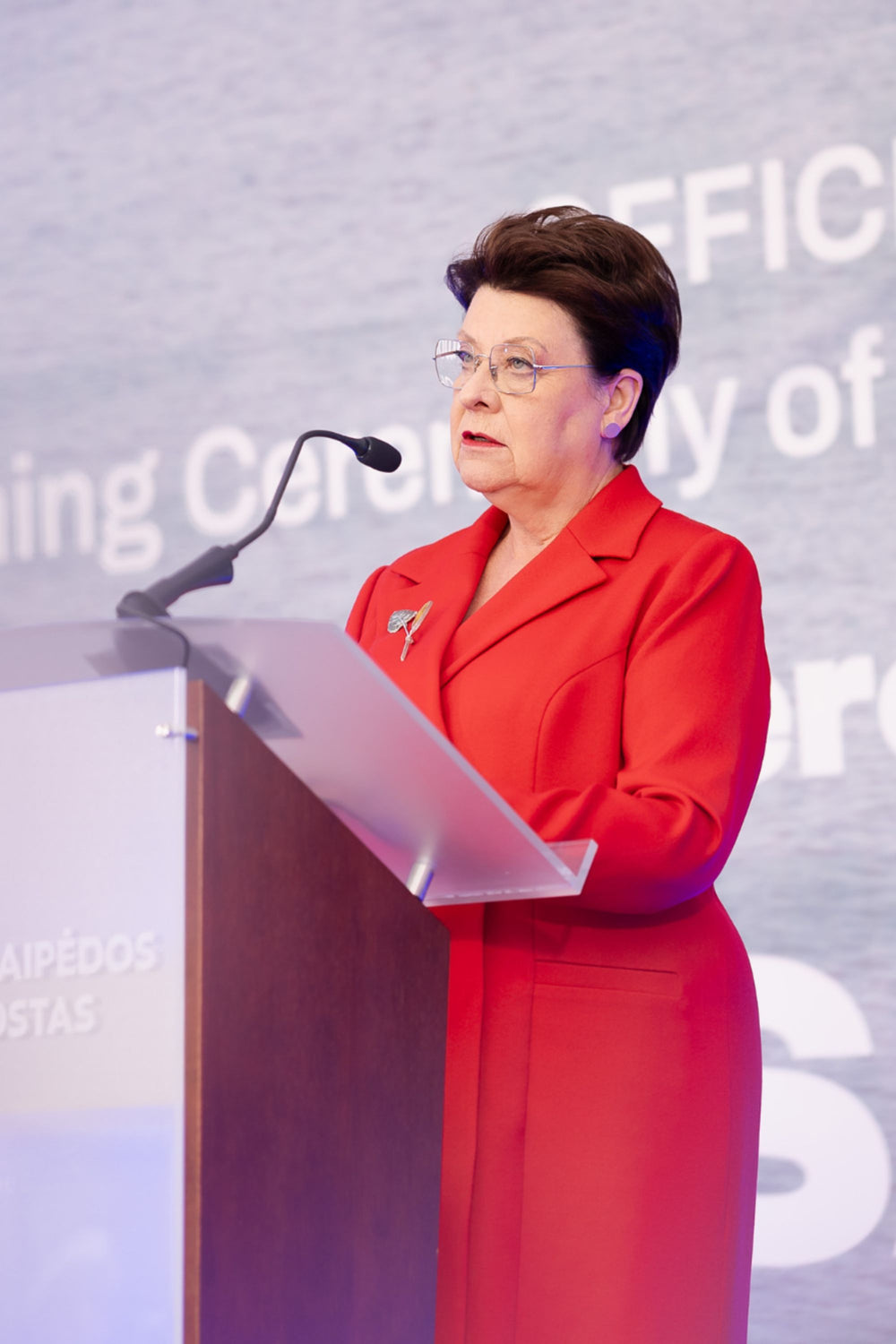 Elegant woman in a luxurious red suit standing at a podium with a microphone, against a backdrop with text.