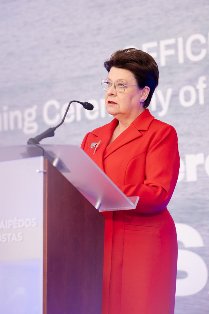 Elegant woman in a luxurious red suit standing at a podium with a microphone, against a backdrop with text.