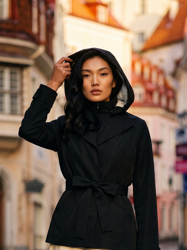 A woman wearing a black hooded trench coat with a tie belt stands on a blurred street with traditional European buildings in the background.