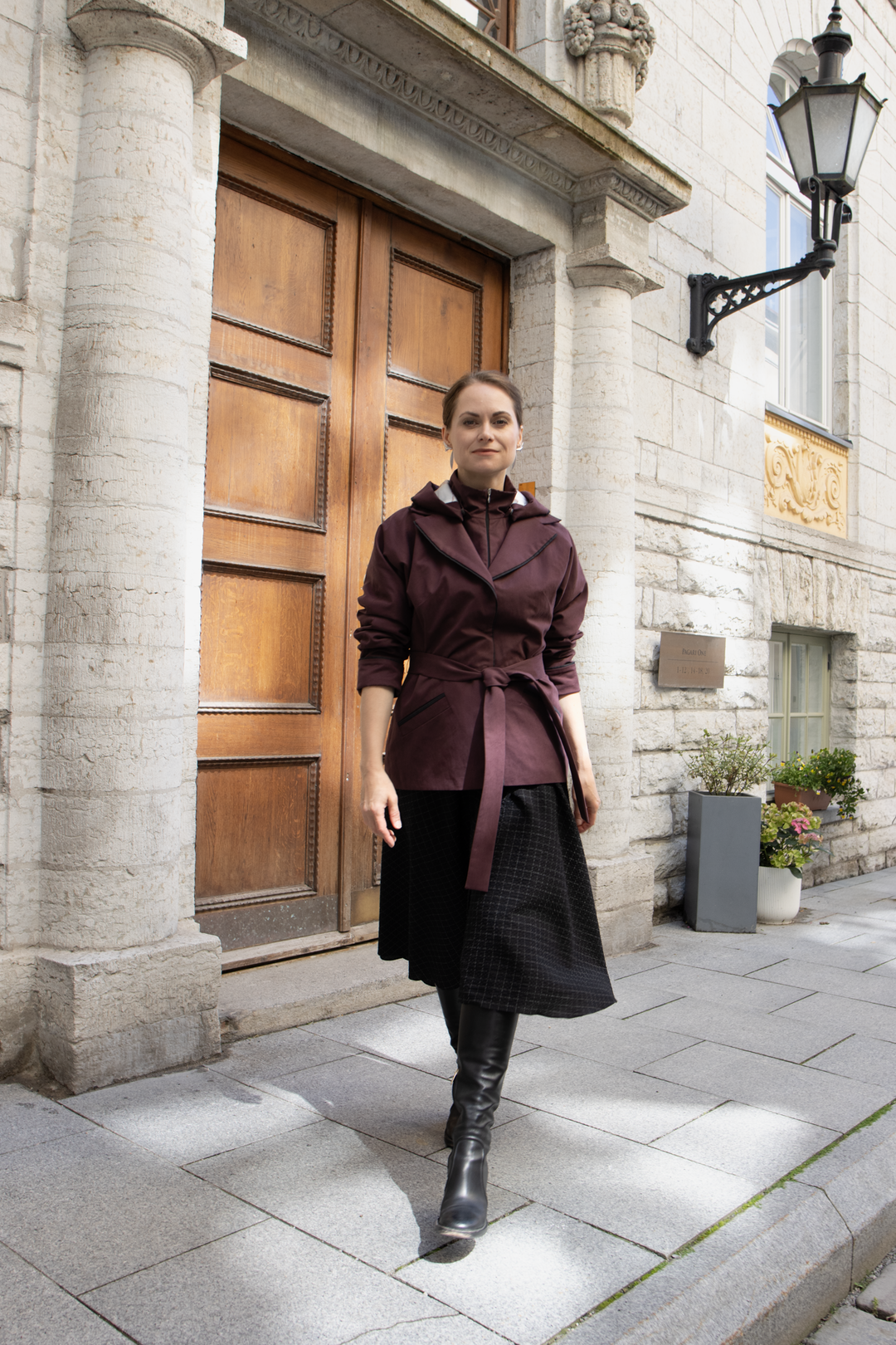 A woman wearing a dark burgundy wrap trench coat with a tie waist, a black skirt, and black boots stands on a city street