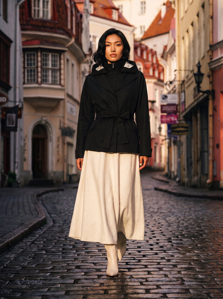 A woman stands on a cobblestone street wearing a black belted jacket with a high collar and a long cream skirt, photographed in a narrow European-style alley with buildings on both sides.