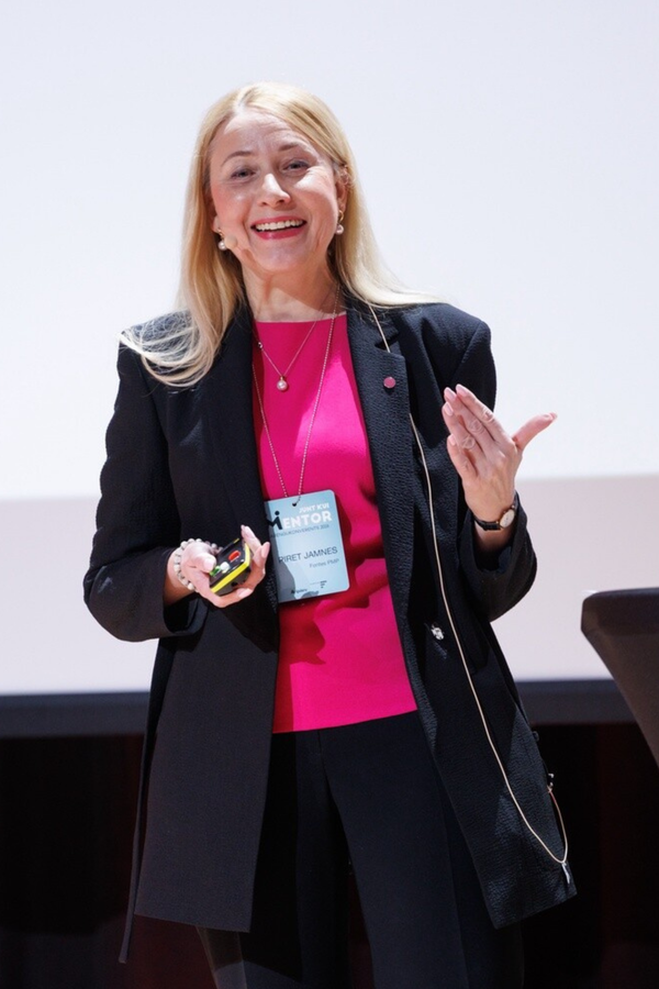 woman in a bright fuchsia top and balck jacket is speaking on stage.
